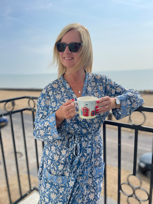 Michelle wearing a blue and white cotton robe from Happy Cabbage holding a beach theme mug and enjoying the beachside seaview at Littlestone in Kent
