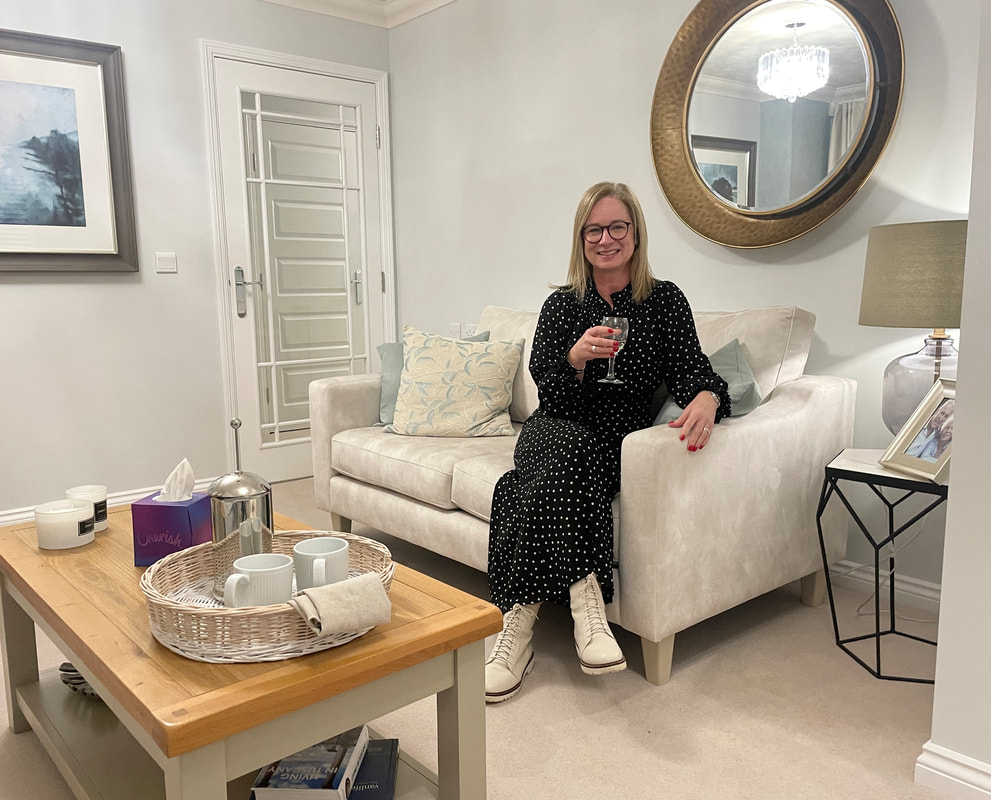 Michelle is sitting on a cream sofa drinking wine at beeches lodge, churchill retirement living