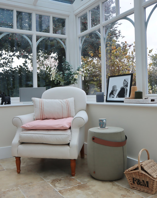 a corner of the conservatory, with a cream arm chair, a sage green footstool and a F&M basket