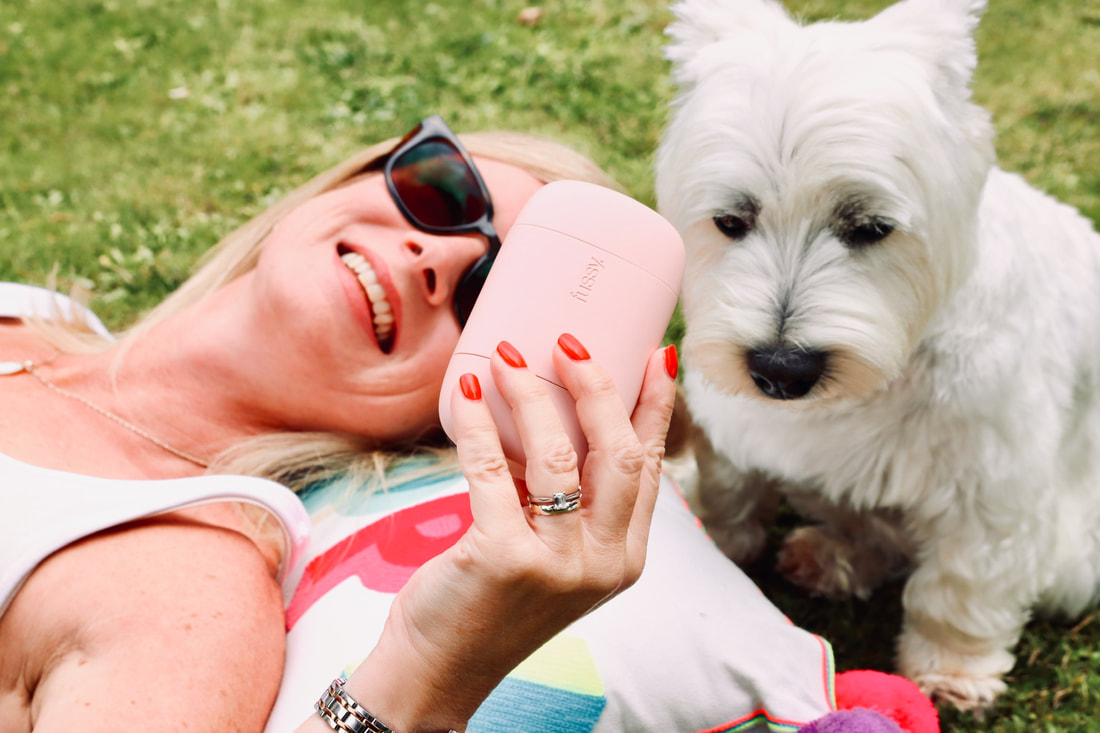 Michelle is laying on the grass with her westie | holding blush pink case fussy deodorant