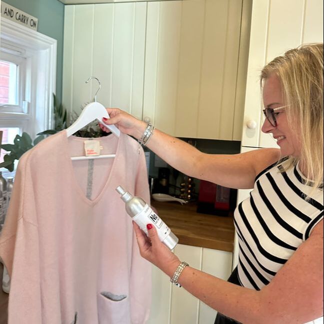 Michelle is wearing a stripe top and standing in her laundry room using the knitwear mist from clothes doctor, holding a pink cashmere jumper