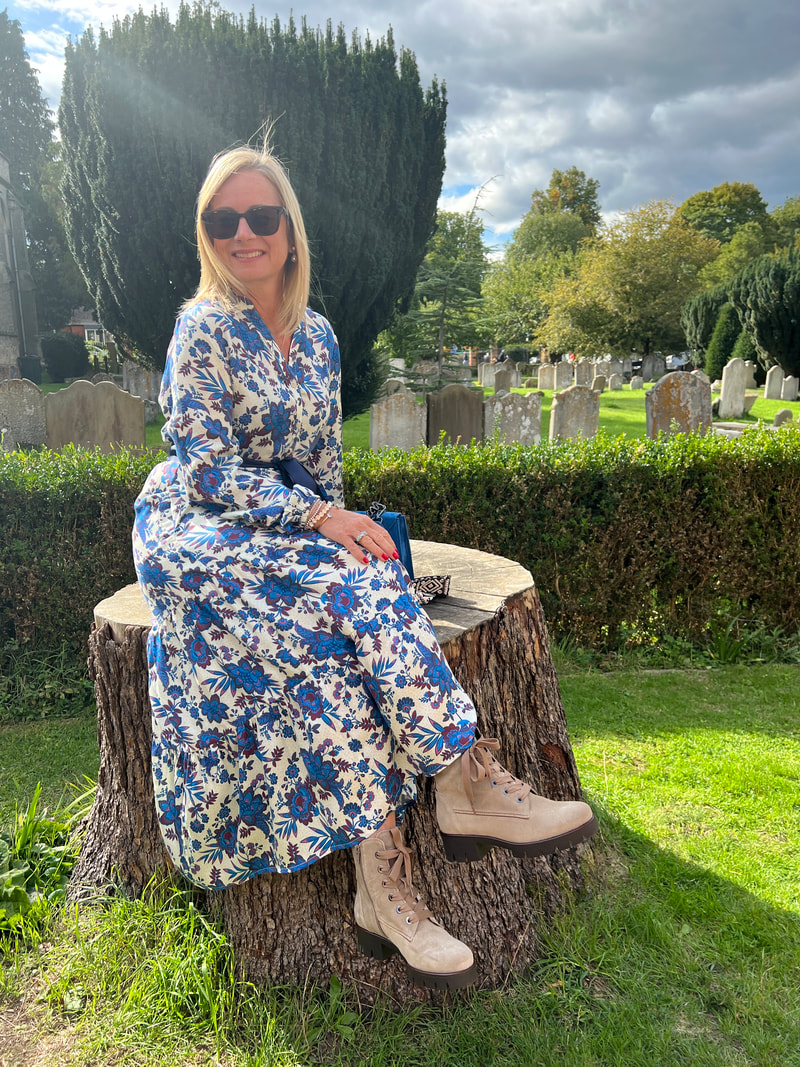 Michelle is sitting on a large tree stump in the church yard at all saints in marlow and is wearing a blue dress and desert boots