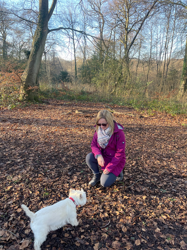 michelle is kneeling down to greet her westie dog  in the woods near marlow.
