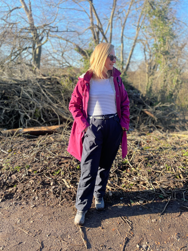 Michelle is standing in the woods with blue skies in the winter wearing waterproof trousers from cotton traders and a cherry pink waterproof coat