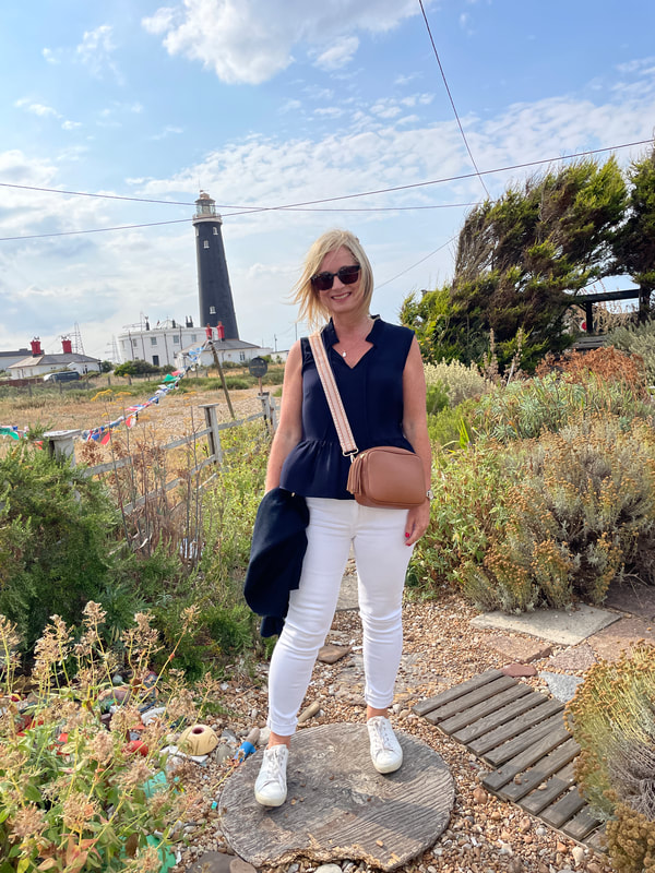 Michelle standing in the garden at the art gallery in Dungeness wearing white jeans and blue top with the old lighthouse behind