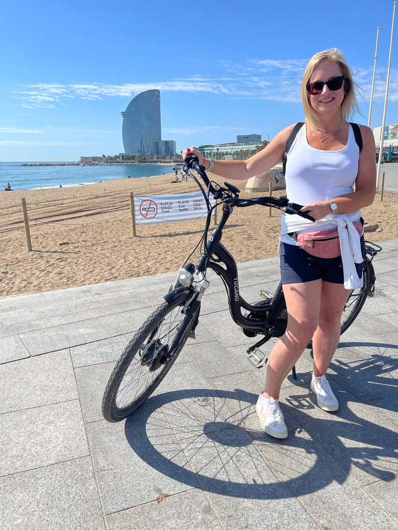 woman in shorts and white top standing by an e-bike at Barcelona beach with W hotel in background