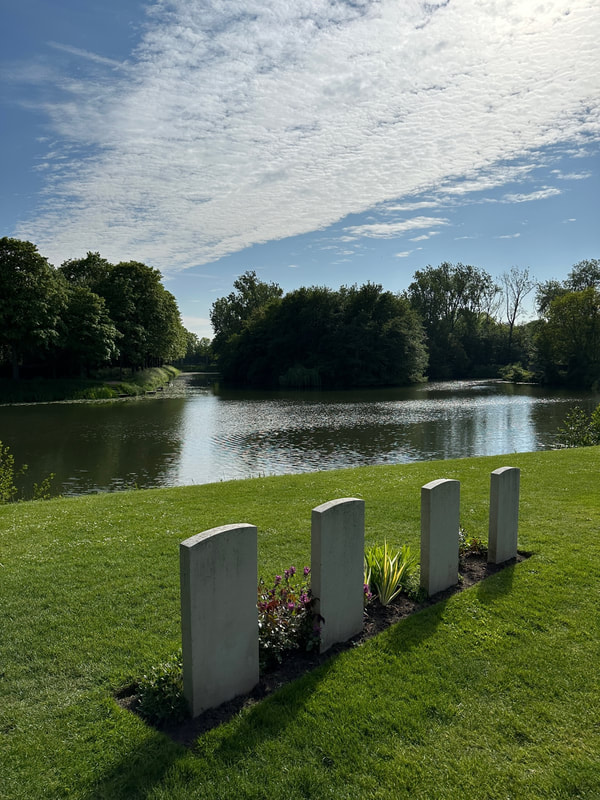 a british war cemetery in ypres showing four white headstones on the grass overlooking the river, the sun is reflecting on the water, it is very peaceful