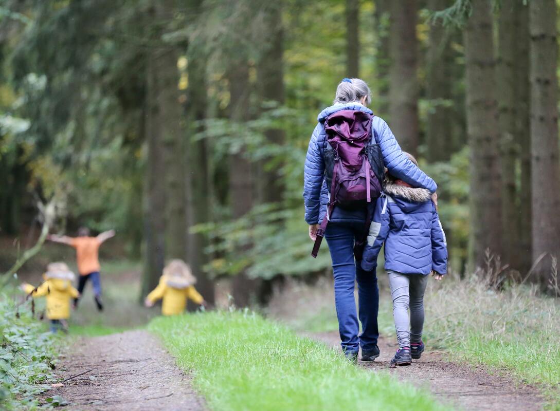 a woman is walking in the forest wearing a blue jacket and a backpack, she has her arm around a child and there are three other children playing around her.