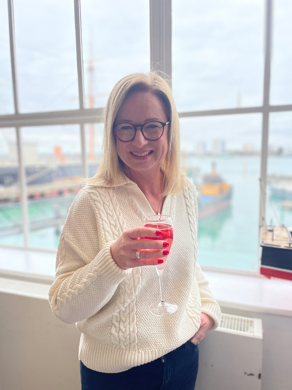 Michelle is drinking a pink kia royale, wearing a cream jumper and standing by the window at boathouse 4 in Portsmouth Historic Dockyard
