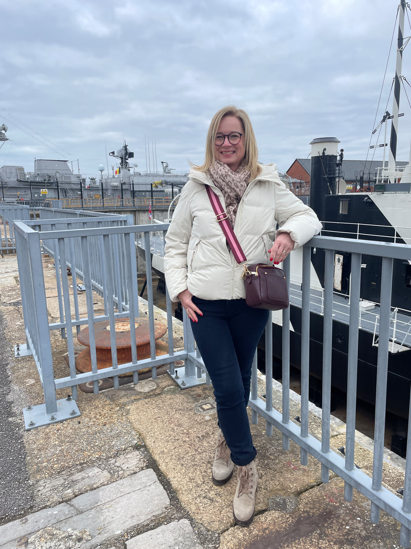 Michelle is standing by grey railings in front of a naval ship at Portsmouth Historic Dockyard