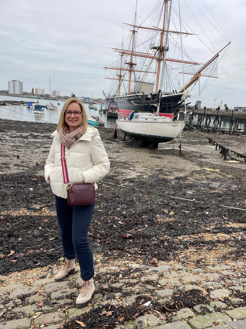Michelle is standing on the beach area at Portsmouth waterfront, there is an historic ship behind her.