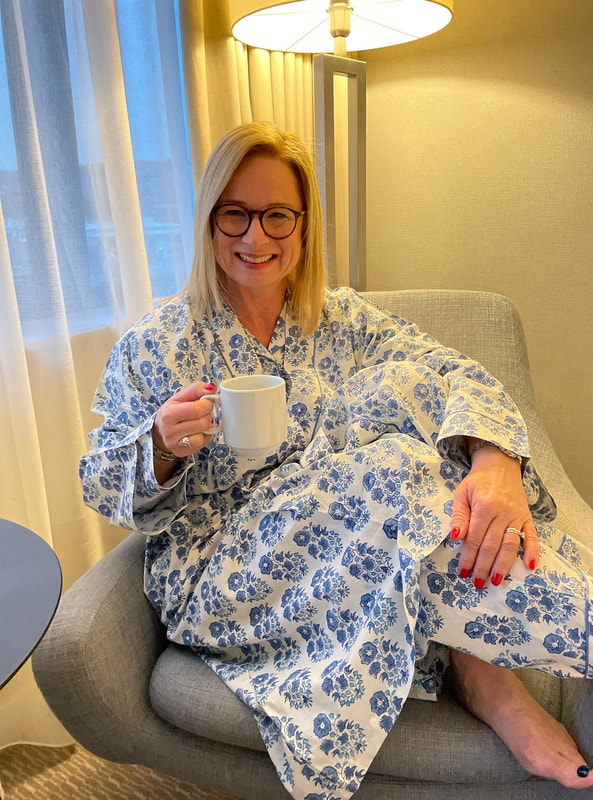 Michelle is sitting on a grey armchair wearing blue and white cotton robe from Rooh London, holding a cup of tea