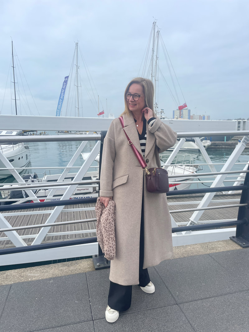 Michelle is wearing a neutral long coat and a burgundy cross body bag and is standing by the waterfront at Portsmouth