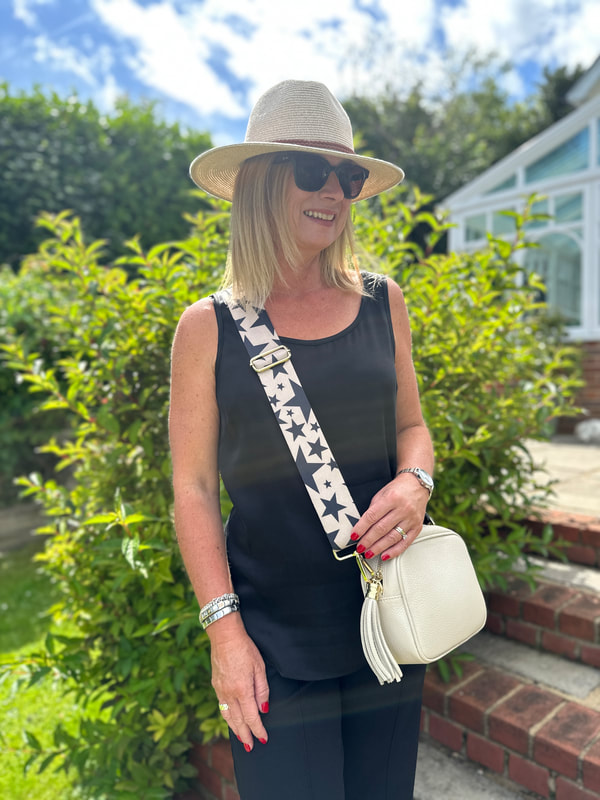 Michelle is in the garden and wearing a black vest top and black trousers with a cream cross body bag and cream hat