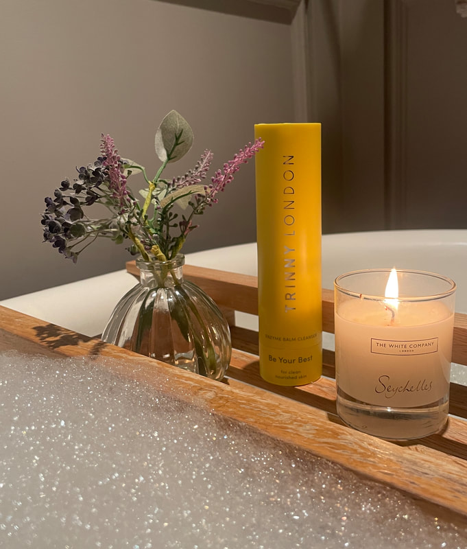 Trinny London Be Your Best Enzyme Balm Cleanser on a bath board with a candle, flowers and bubble bath
