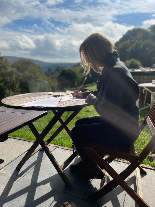 michelle is sat at a wooden table writing in a notebook with the view of the tintern hills in front of her