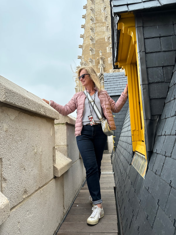 michelle is wearing blue jeans and a pink jacket and standing by a yellow frame window on the roof at the belfry, in flanders fields museum