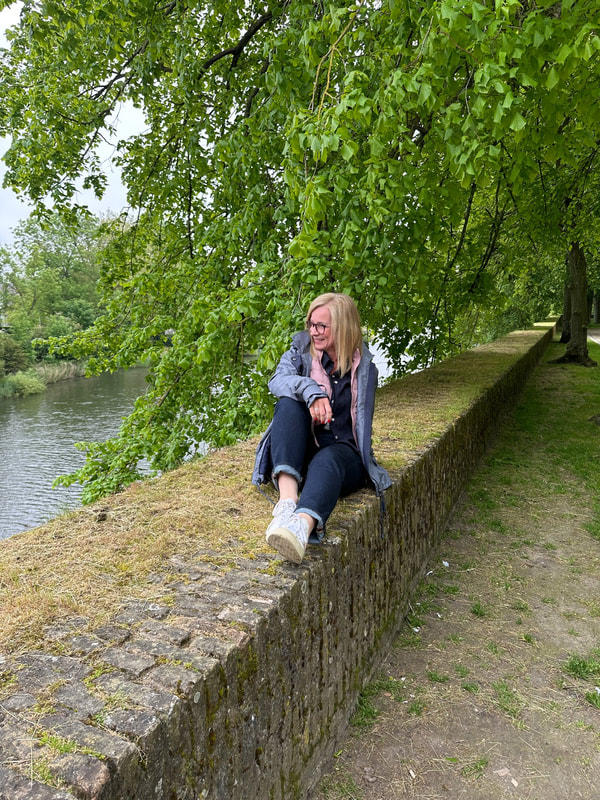 Michelle is sitting on a wall by the river at Ypres, she is wearing blue jeans and a grey jacket